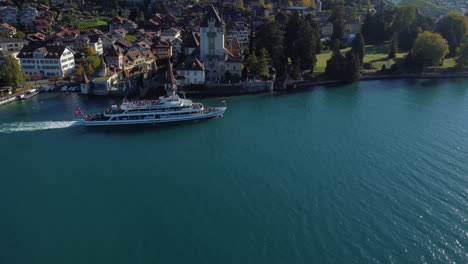 Toma-Aérea-De-Seguimiento-De-Un-Barco-De-Pasajeros-Y-Vista-Del-Castillo-De-Oberhofen-En-Suiza,-En-Un-Día-Soleado