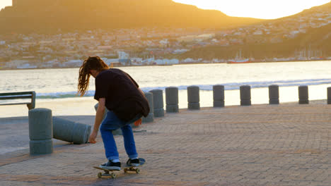 Rear-view-of-young-caucasian-man-jumping-with-skateboard-on-beach-promenade-during-sunset-4k