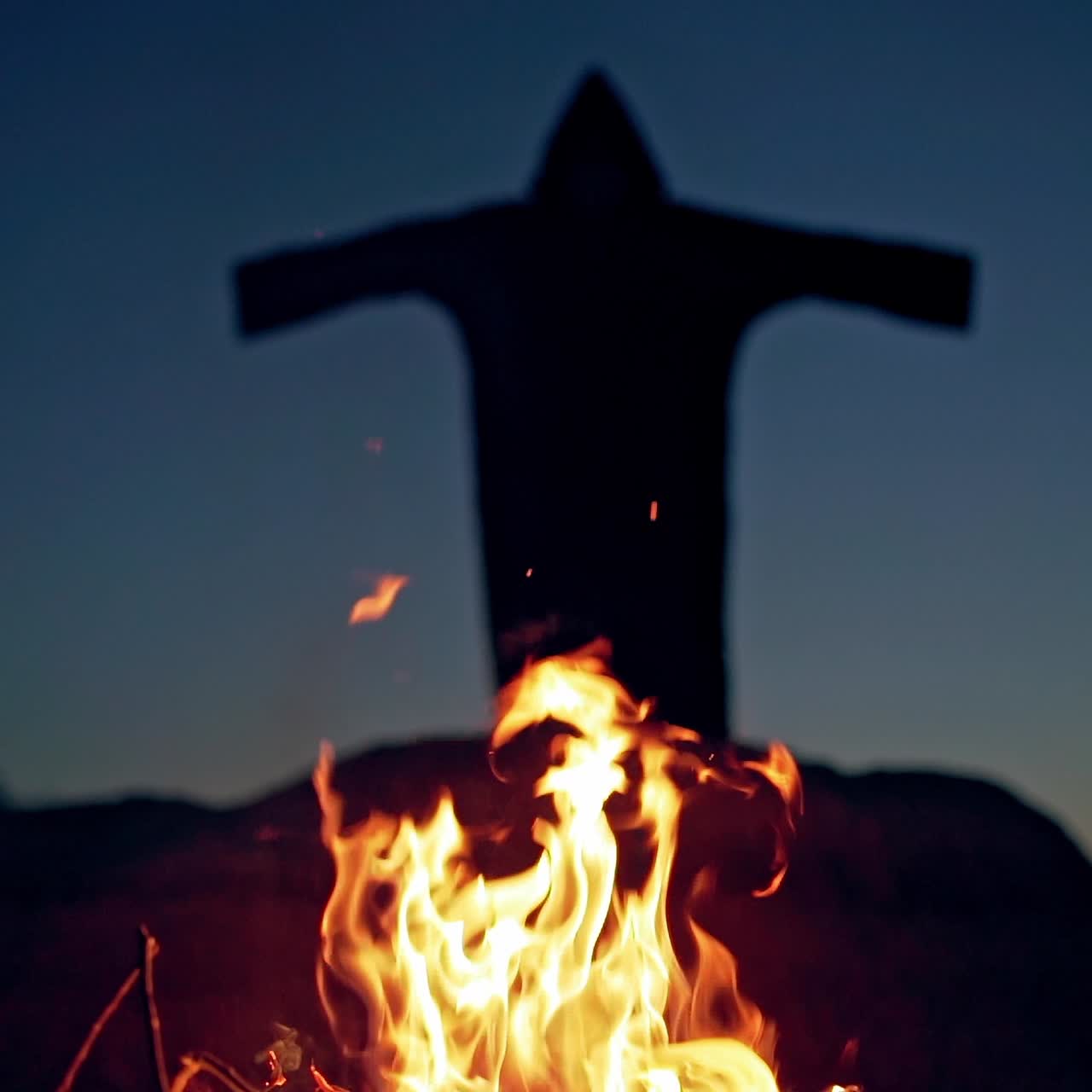 Scary silhouette od black figure on the hill behind fire. Orange flame at night and horrorful death in black cloak with hood is waving arms. Halloween concept.