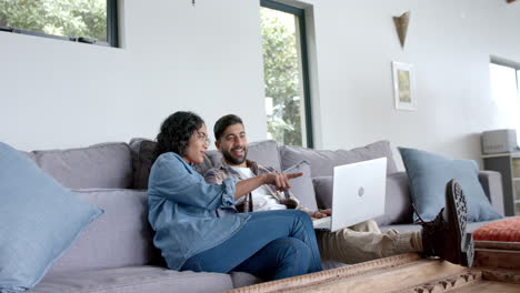 happy biracial couple sitting on sofa and using laptop at home, in slow motion