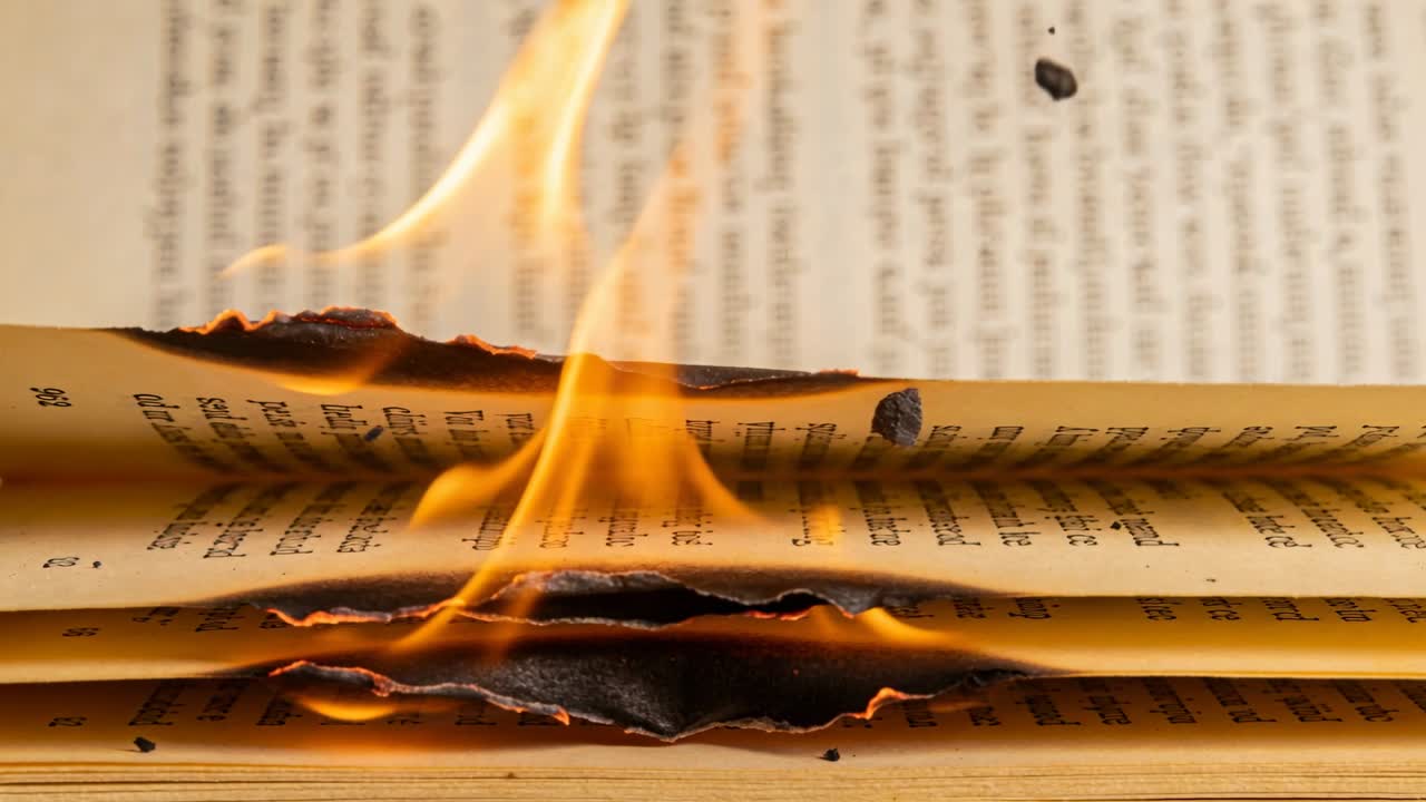 Pagine brucianti di un libro aperto che si arricciano e lasciano cadere braci incandescenti su un tavolo di legno, con i bordi carbonizzati.