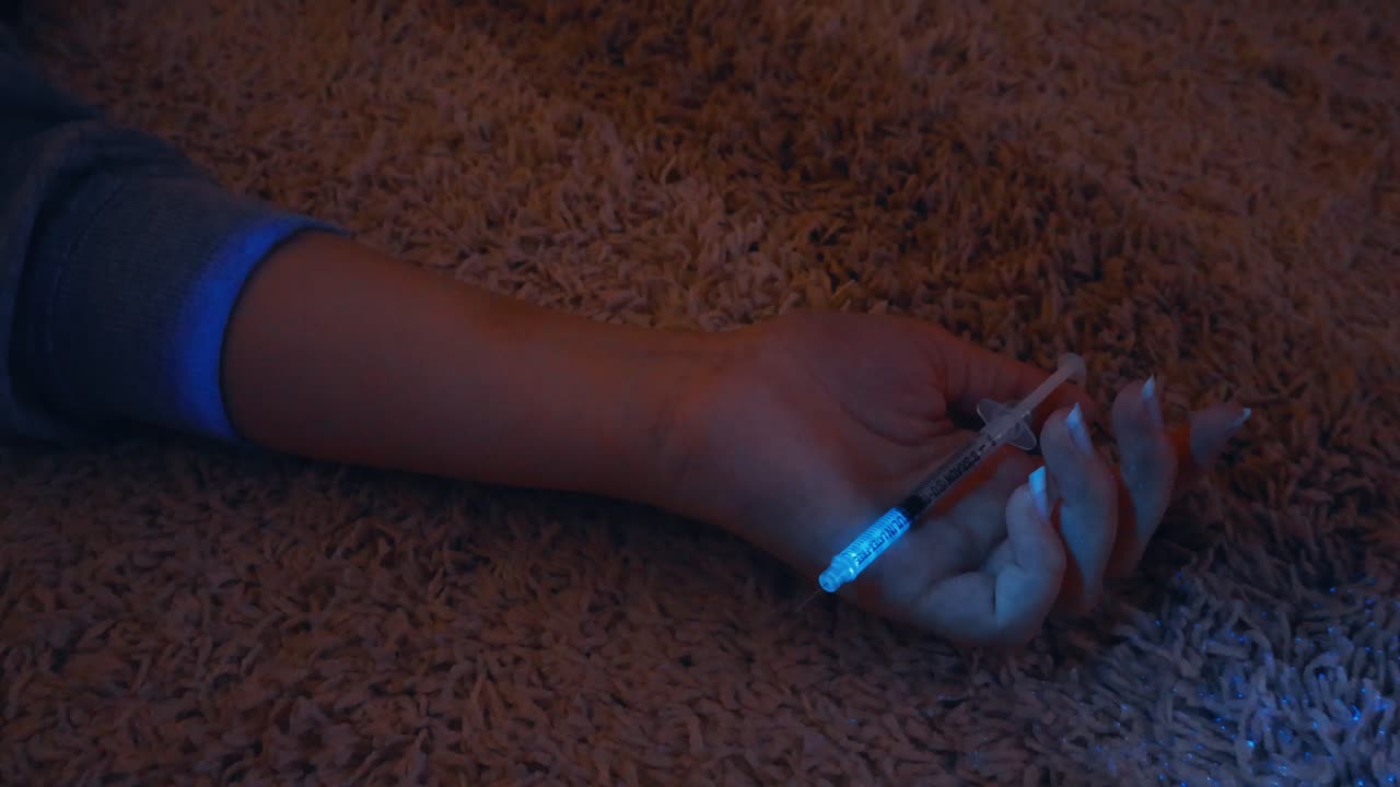 Ultraviolet light beam scanning a woman's arm holding a syringe on the floor of a crime scene, revealing forensic evidence of a possible drug overdose, suicide, or murder mystery
