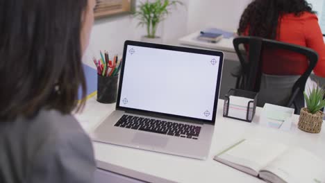 mujer de negocios biracial sentada en un escritorio, usando una computadora portátil con espacio de copia en una oficina moderna