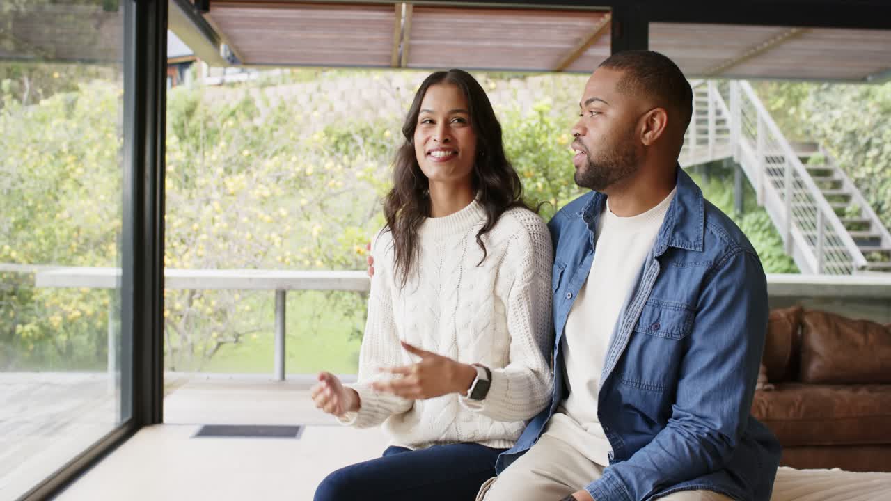 Diverse couple gazing at garden view through window at home, woman describing and man nodding
