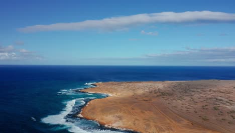 a wide arid peninsula curves into rolling blue waves, blending golden sand and rugged coastline beneath a brilliant sky with drifting white clouds.