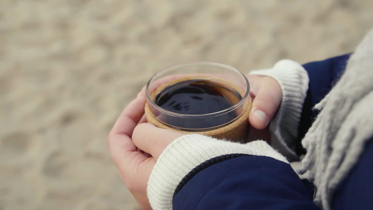 추운 날 해변에서 뜨거운 커피 머그잔을 손에 들고 몸을 따뜻하게 하는 여자