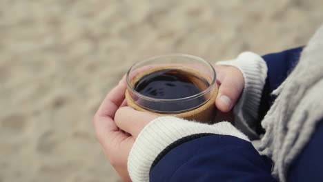 Mujer-Sosteniendo-Una-Taza-De-Café-Caliente-En-Sus-Manos-En-Una-Playa-En-Un-Día-Frío-Para-Calentarse