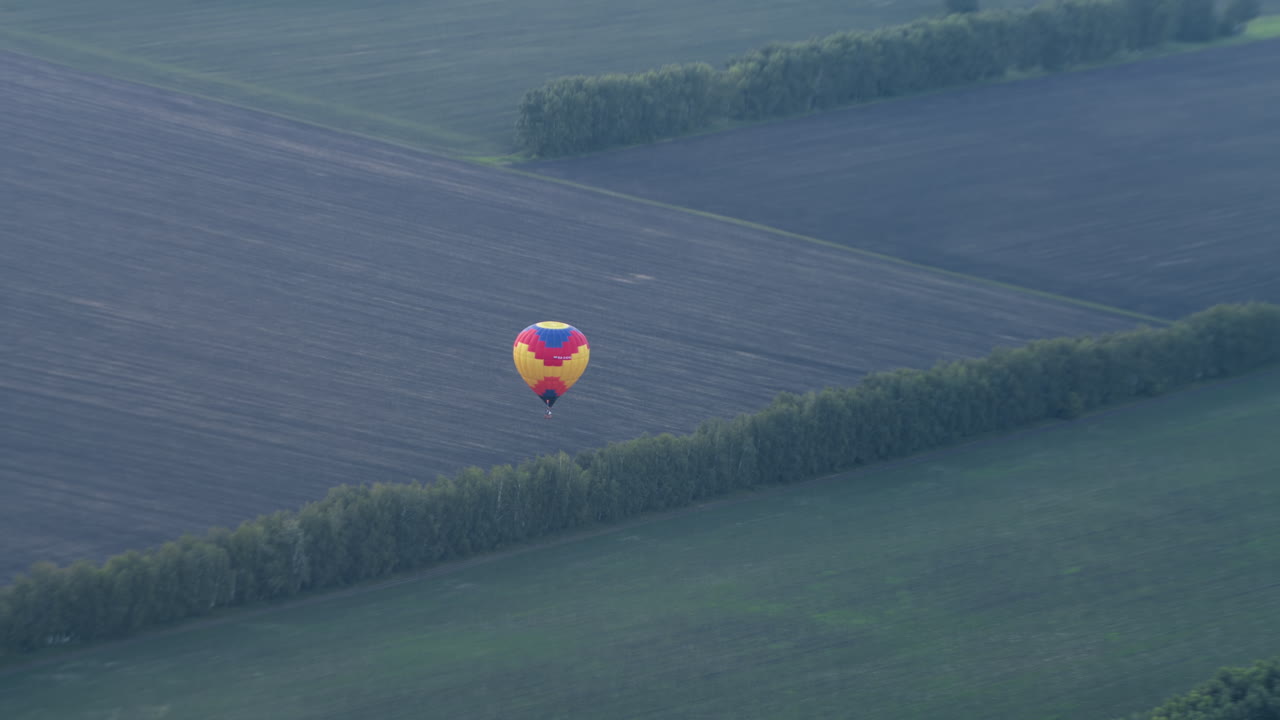 Distant view of colourful hot air balloon descending over sprawling tilled fields bordered by lush tree lines, wicker basket swinging beneath rippled canopy