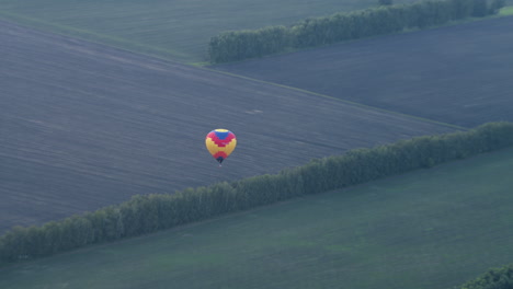 distant view of colourful hot air balloon descending over sprawling tilled fields bordered by lush tree lines, wicker basket swinging beneath rippled canopy