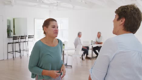 Two-senior-female-friends-standing-together-socialising-before-their-ballroom-dancing-class