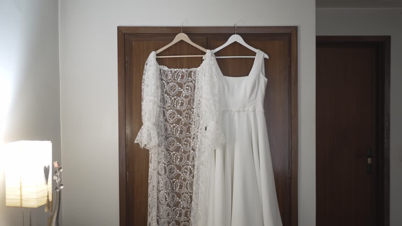 Abito da sposa bianco e giacca bolero in pizzo appesi a grucce contro una porta di legno in una luminosa stanza interna.