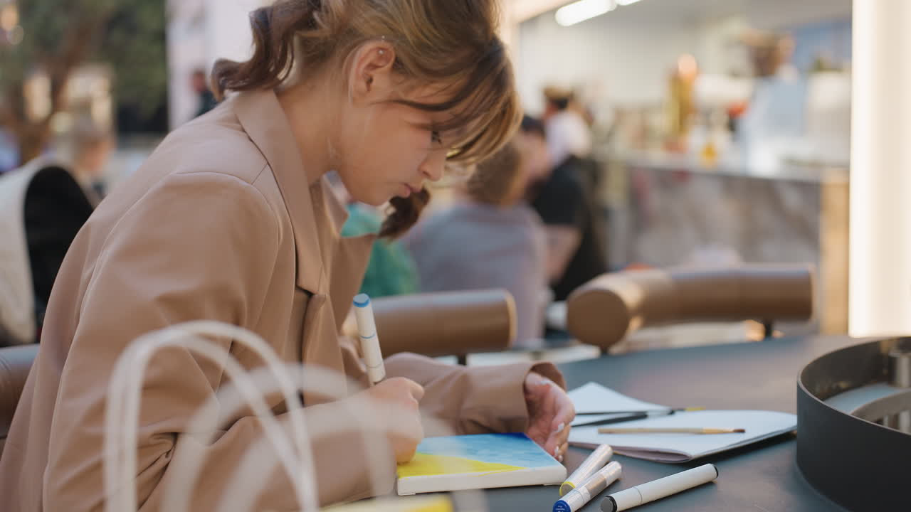 Молодая женщина пишет что-то в блокноте в кафе, на ней бежевое пальто и приспущенная маска, рядом с ноутбуком — стикеры и ручка, сосредоточенное выражение лица, городской фон с боке, планирует расписание и делает наброски идей, теплые тона