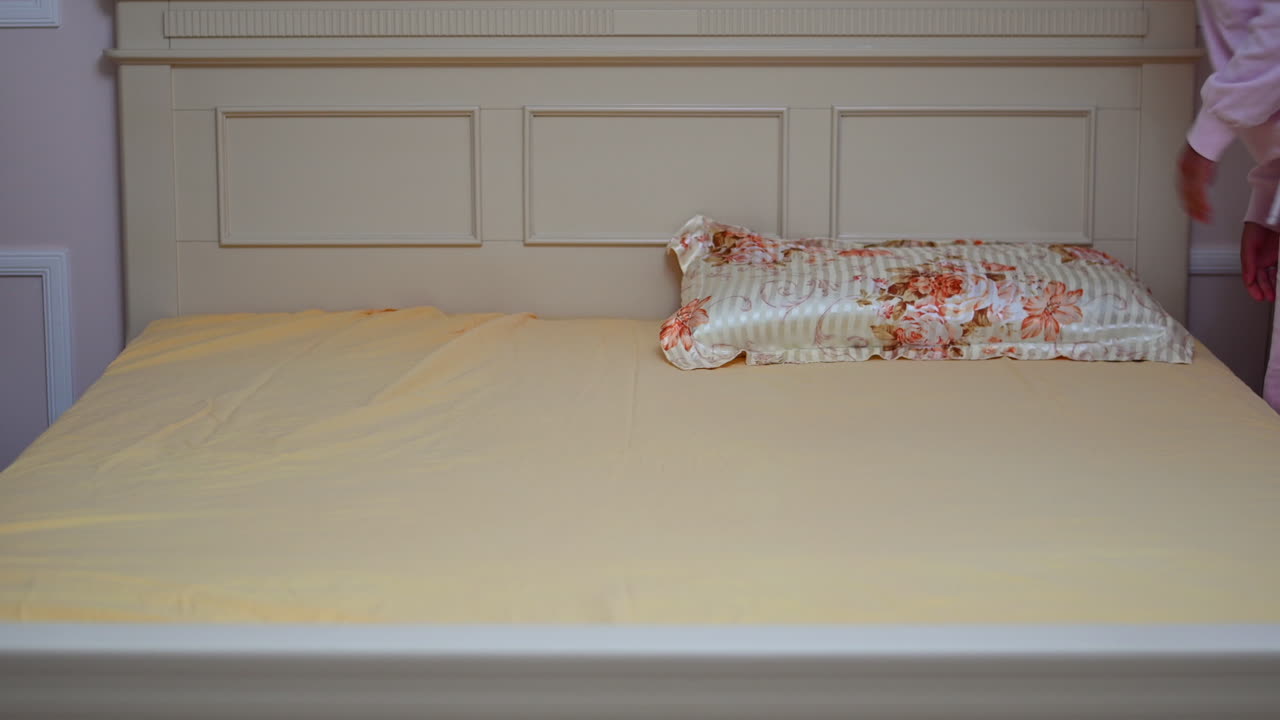 A person is making a bed with a yellow sheet, tucking it neatly into the corners for a clean and organized appearance. The soft fabric complements the light-colored bedroom