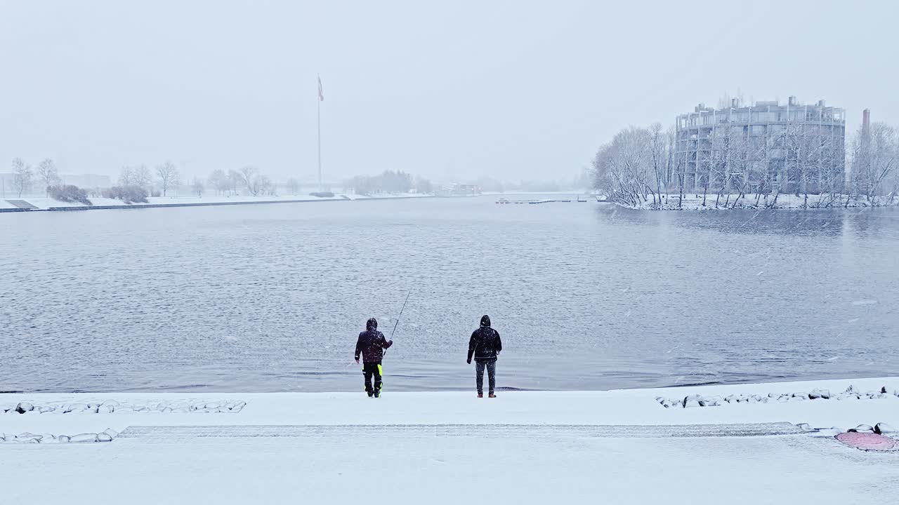 Snowy riverside moment as two people quietly fish during winter in Latvia