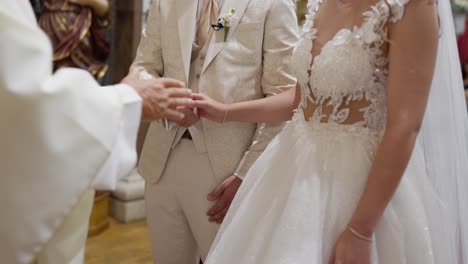primer plano de una ceremonia de boda donde un sacerdote oficia el intercambio de votos entre una pareja vestida con elegantes trajes blancos