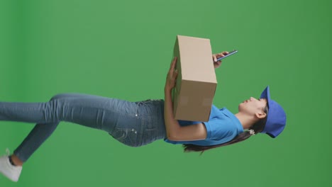 side view of asian female courier in blue uniform walking and using smartphone while delivering a carton on green screen background in the studio