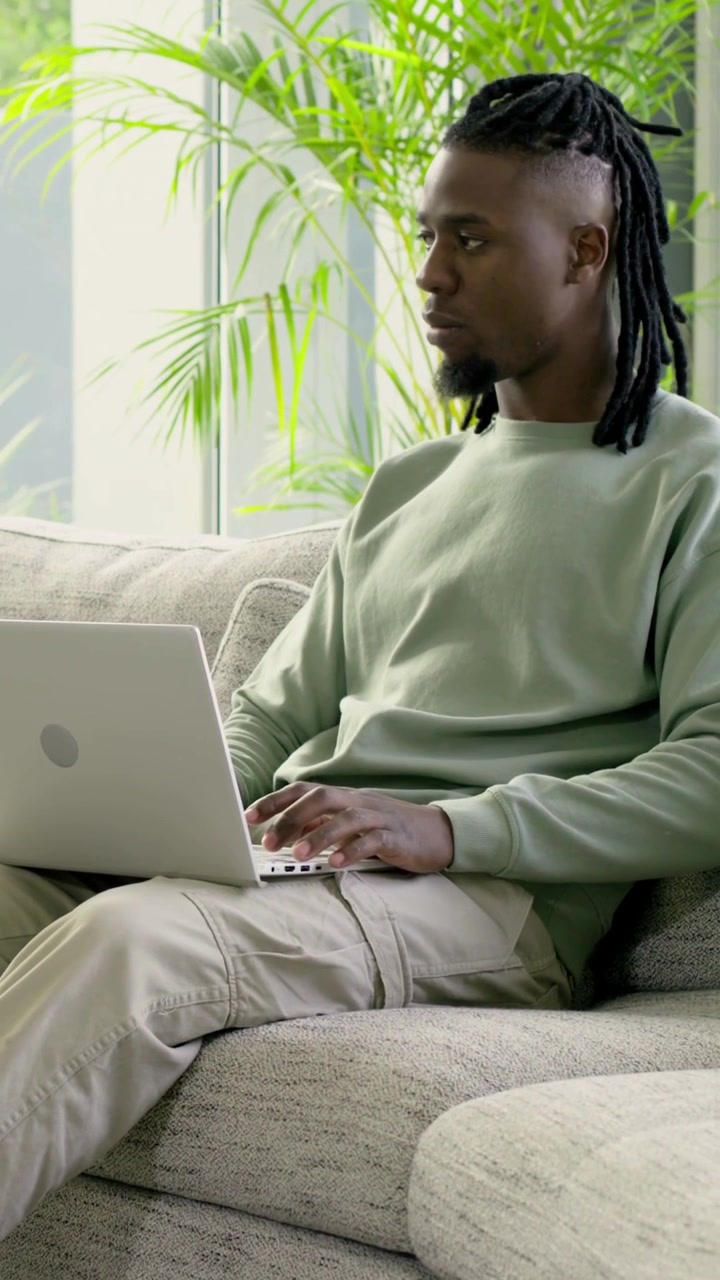 Vertical video: Typing man on laptop in living room pausing to gaze at window as wind swaying plant