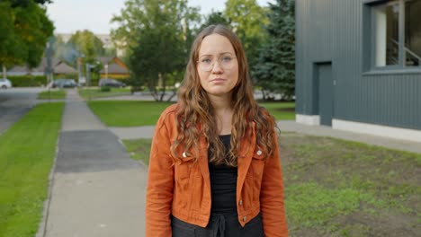 -Woman-in-Orange-Jacket-Stands-on-Sidewalk,-Gazing-Away-with-a-Somber-Expression-Outside-an-Office