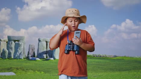 asian boy with a hat and binoculars looking at smartphone then saying wow. boy researcher examines something while traveling in stonehenge, travel tourism adventure concept