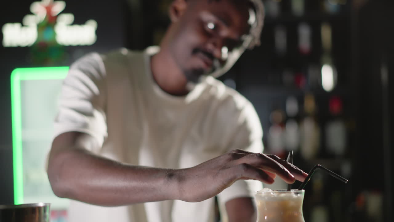 Bartender adding final touches to cocktail, placing straw in drink with focus. Behind, illuminated Heineken neon sign and bar shelves filled with liquor bottles, enhancing vibrant bar scene