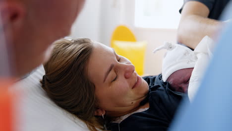 woman is crying from happiness to hold her just born baby. caucasian mom lies in bed holding a newborn on her chest and husbands sits beside.