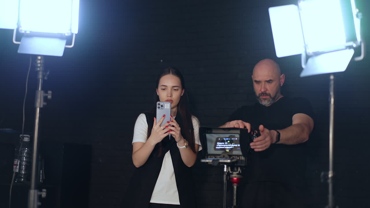 Man and young woman taking video in the studio. Girl uses a phone and man uses a professional camera. Backstage work.