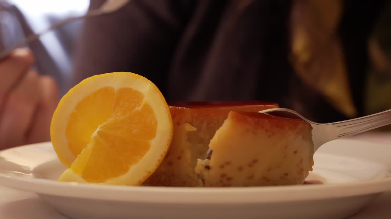 Spoon cutting into caramel flan with orange slice on white plate
