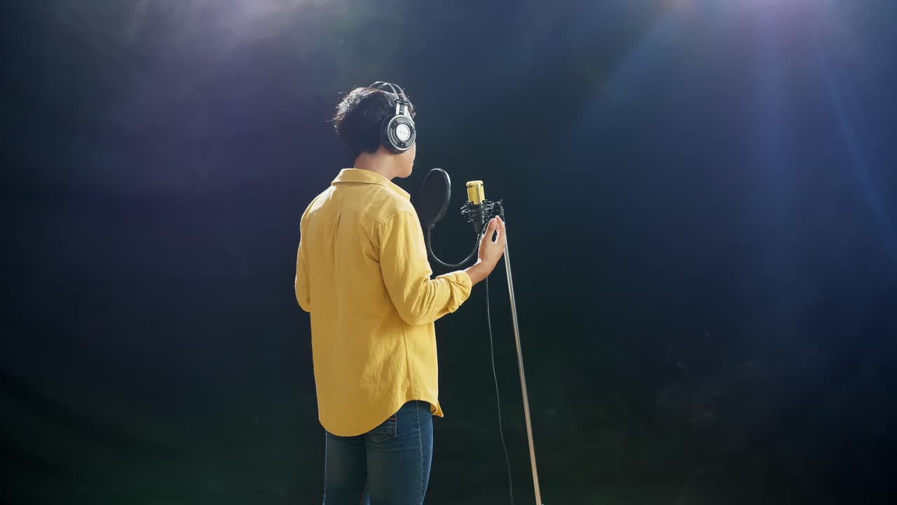 Rückansicht eines jungen Jungen mit Kopfhörern, der in ein Kondensatormikrofon singt, vor weißem Rauch auf schwarzem Hintergrund