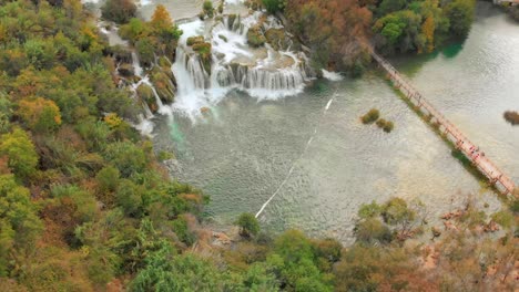 a drone captures the krka waterfalls in stunning detail. people cross a wooden bridge in the foreground, adding scale and perspective.