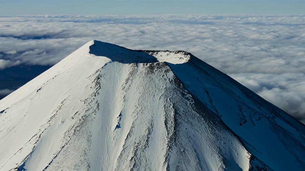 Un cono vulcanico innevato si erge sopra una densa coltre di nuvole mentre il punto di osservazione si allarga gradualmente, rivelando i contorni frastagliati della cima, i fianchi in ombra e la neve luminosa baciata dal sole sotto un cielo azzurro.