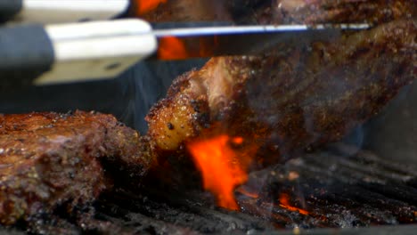 Un-Par-De-Pinzas-Para-Carne-Convierten-Un-Jugoso-Filete-De-Costilla-Casi-Cocido-En-Una-Parrilla-Mientras-Las-Llamas-Anaranjadas-Se-Disparan-Desde-Abajo-En-Cámara-Lenta