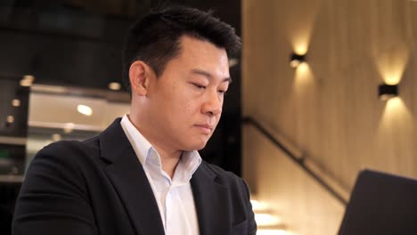 serious asian businessman is typing message checking info using laptop in business center lobby. portrait of focused man wearing formal black suit standing in hall with computer. an urgent matter.