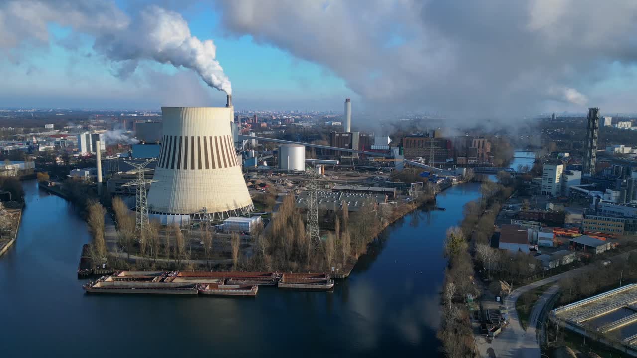 Aerial view of a thermal power plant emitting smoke on a sunny day, with a river and a city in the background. Spectacular aerial view flight speed ramp hyper motion time lapse