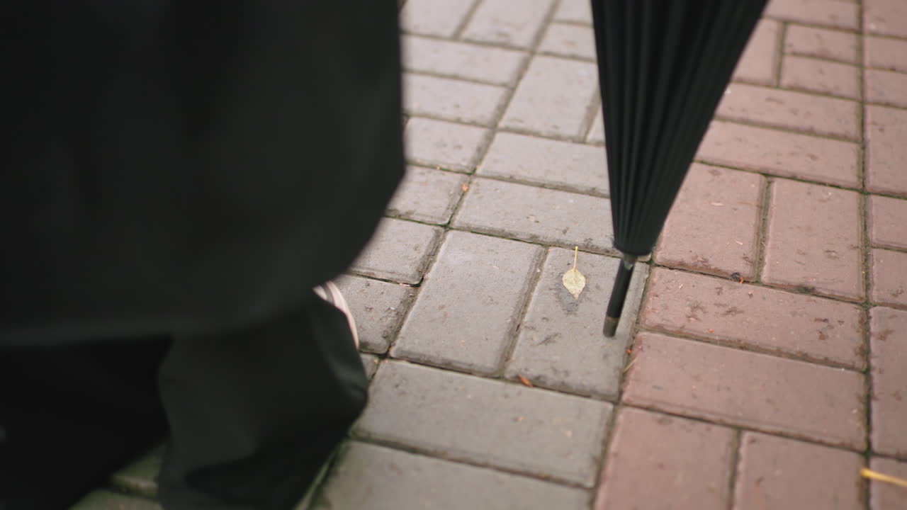 Primo piano di una persona con un cappotto nero, in piedi su un marciapiede lastricato, che indossa scarpe da ginnastica bianche e tiene un ombrello nero chiuso, a simboleggiare attesa, preparazione, protezione dagli agenti atmosferici, resilienza, sicurezza personale