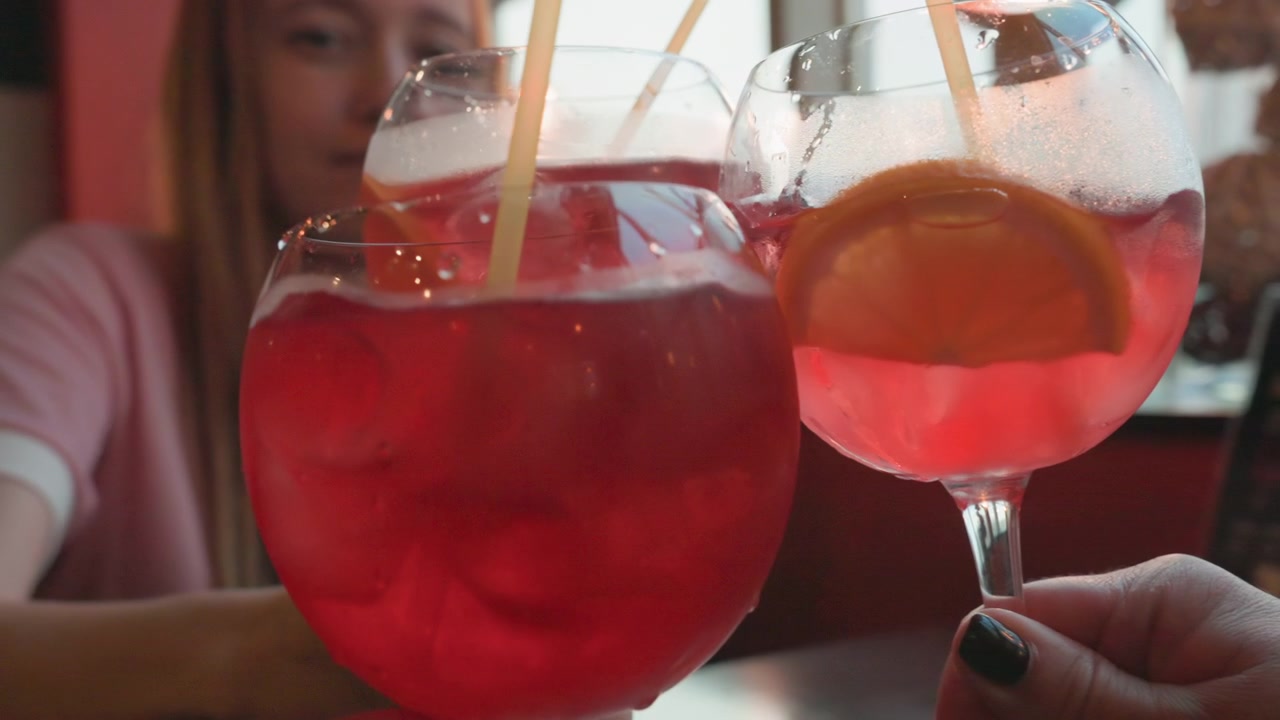 Giovane donna tiene un grande bicchiere di bevanda rossa con ghiaccio e cannuccia; è visibile anche la mano di un'altra donna che tiene bevande simili. L'ambiente sembra essere un caffè o un bar al chiuso vicino a finestre con luce soffusa.