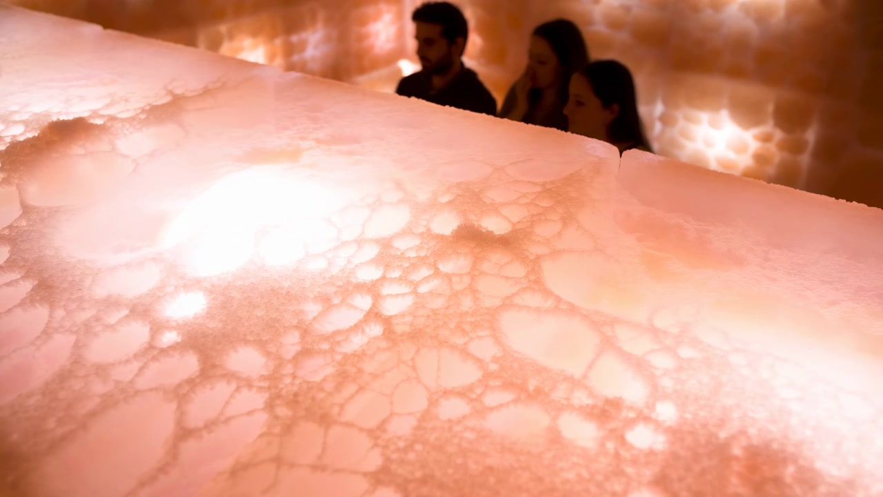 Una calda illuminazione si diffonde attraverso un'ampia superficie traslucida e testurizzata in primo piano, mentre tre persone rimangono dietro di essa, compiendo piccoli movimenti all'interno di uno spazio chiuso e delicatamente illuminato.