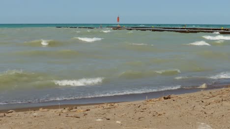 Las-Olas-Se-Estrellan-Suavemente-En-Una-Playa-De-Arena-De-Izquierda-A-Derecha,-En-Un-Día-Soleado,-Con-Vistas-A-Un-Par-De-Muelles