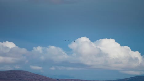 Una-Pequeña-Bandada-De-Pájaros-Vuela-Rápido-Sobre-El-Paisaje-Montañoso-Y-Las-Nubes-Al-Fondo