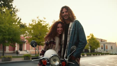 Portrait-of-a-happy-guy-and-girl-with-curly-hair-in-plaid-shirts,-a-girl-sits-on-a-moped,-holds-a-white-helmet-in-her-hands-and-looks-at-her-boyfriend-in-a-denim-jacket-on-the-street-near-the-trees-on-a-wide-road-in-summer