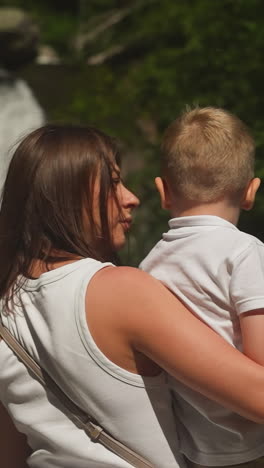 careful mother embraces little boy standing near picturesque waterfall in sunny park. woman takes care of toddler son travelling to river cascade