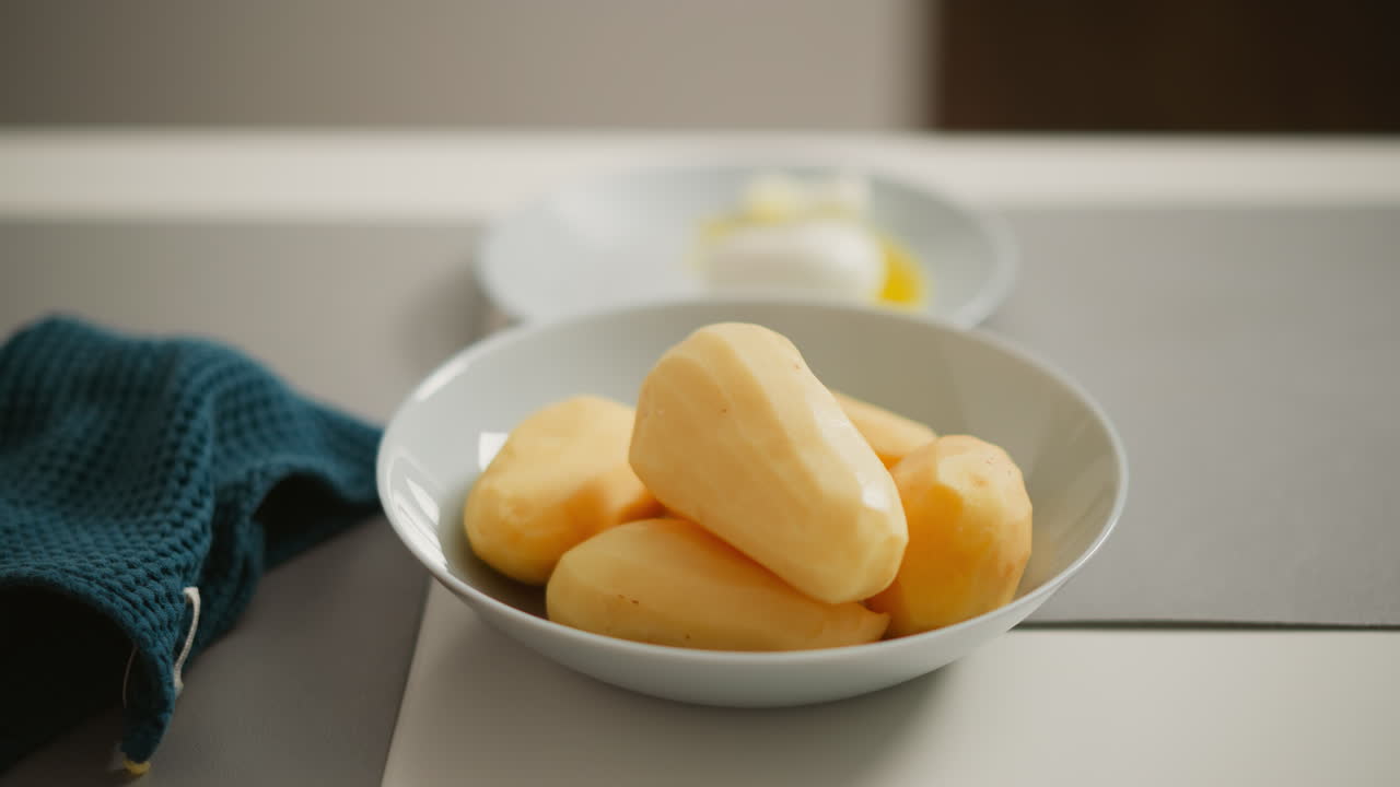 Close up of person dropping peeled potato into white bowl filled with other peeled potatoes on light counter, kitchen rag slightly visible beside plate, soft daylight setting