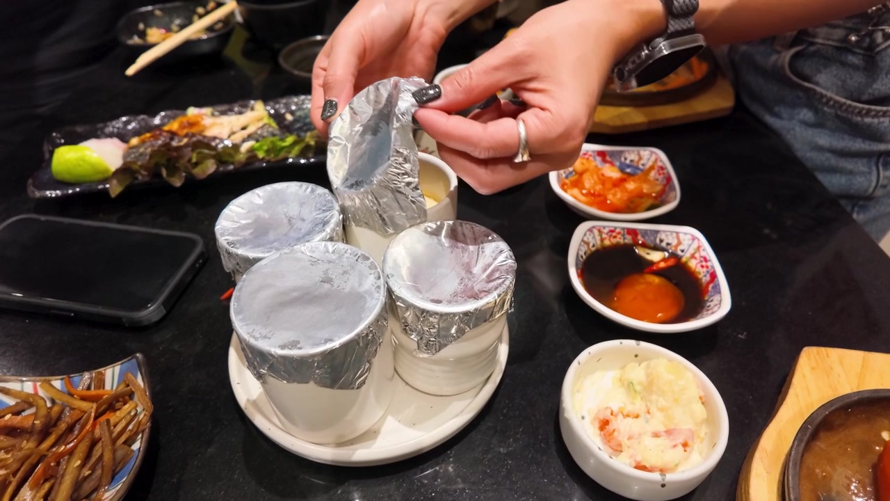 Close-up of hands removing foil from ceramic bowls of steamed egg custard at a restaurant.