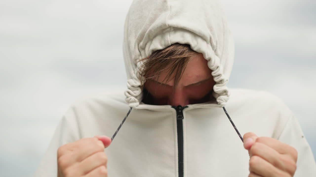 흐린 하늘 아래 후드티를 뒤집어쓰고 조심스럽게 끈을 잡고 있는 젊은 백인 남성, 젖은 머리카락이 이마 위로 덮여 있고, 조심스러운 표정, 시원한 바람, 도시 옥상 배경, 기대감 있는 분위기