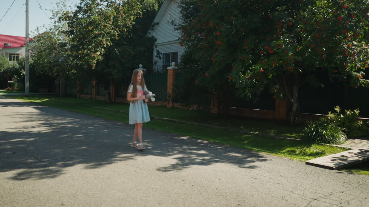 Little girl in light blue dress walks alone on quiet street holding stuffed toy rabbit, casting soft shadow on pavement under scattered tree light in peaceful residential area
