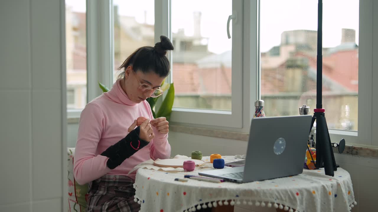 Young Woman with Wrist Brace Doing Embroidery While Watching Laptop