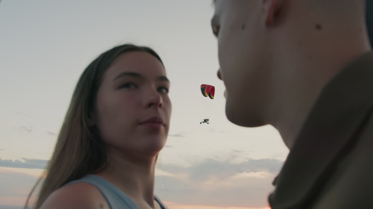Close up of couple facing each other with distant paraglider gliding across pastel sky and soft clouds over patchwork farmland and tree lines creating romantic serene moment at sunset