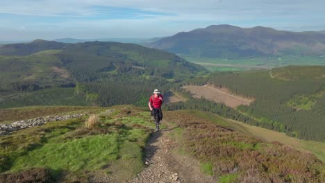 Mountain-walker-trekking-up-path-and-past-camera-on-windy-mountainside-with-view-to-forested-fells-and-green-valley-floor-behind