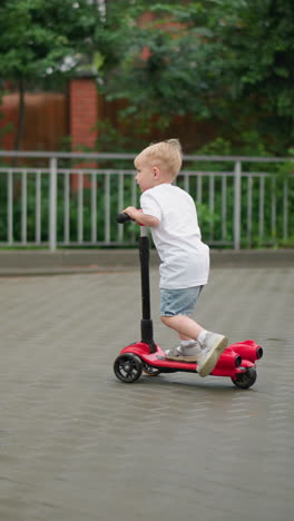 a young child rides a red scooter joyfully down a paved pathway bordered by green grass, metal railings, and a nearby building