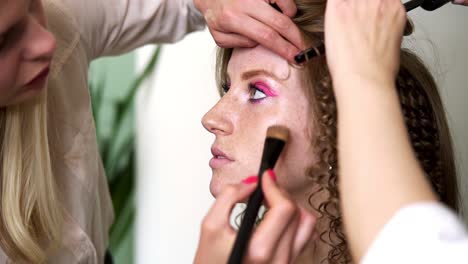 Maquillador-Aplica-Tóner-En-La-Cara-De-Una-Hermosa-Modelo-Con-Pecas.-Maquillador-Trabaja-En-Un-Salón-De-Belleza.-Peluquero-Con-Un-Rizador-Haciendo-Peinados-En-Tándem.-De-Cerca.-Camara-Lenta