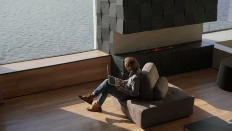 from above wide shot of young man sitting cross-legged on sofa in his luxury apartment with large panoramic windows with sea view, opening pc, working on laptop and thinking